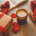 An amber jar candle with a wooden lid placed beside a gift box tied with a red ribbon, surrounded by fresh red roses and scattered petals. An open book adds a cozy, romantic touch.