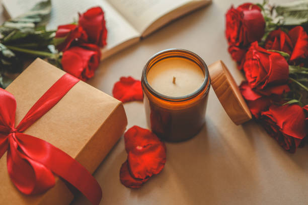 An amber jar candle with a wooden lid placed beside a gift box tied with a red ribbon, surrounded by fresh red roses and scattered petals. An open book adds a cozy, romantic touch.